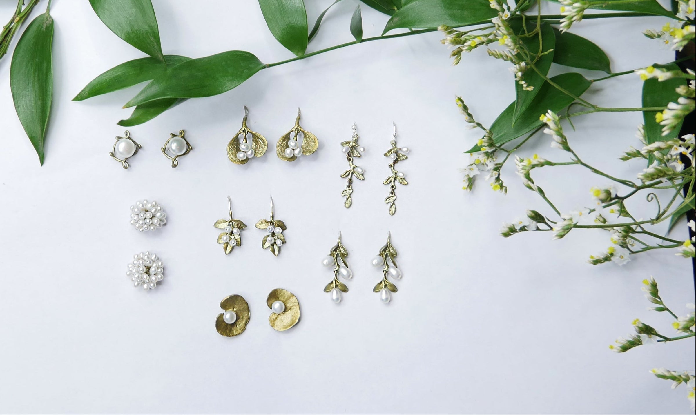 A sheet of white paper displaying various pairs of earrings in gold and silver tones, surrounded by green leaves and white flowers on a wooden surface.