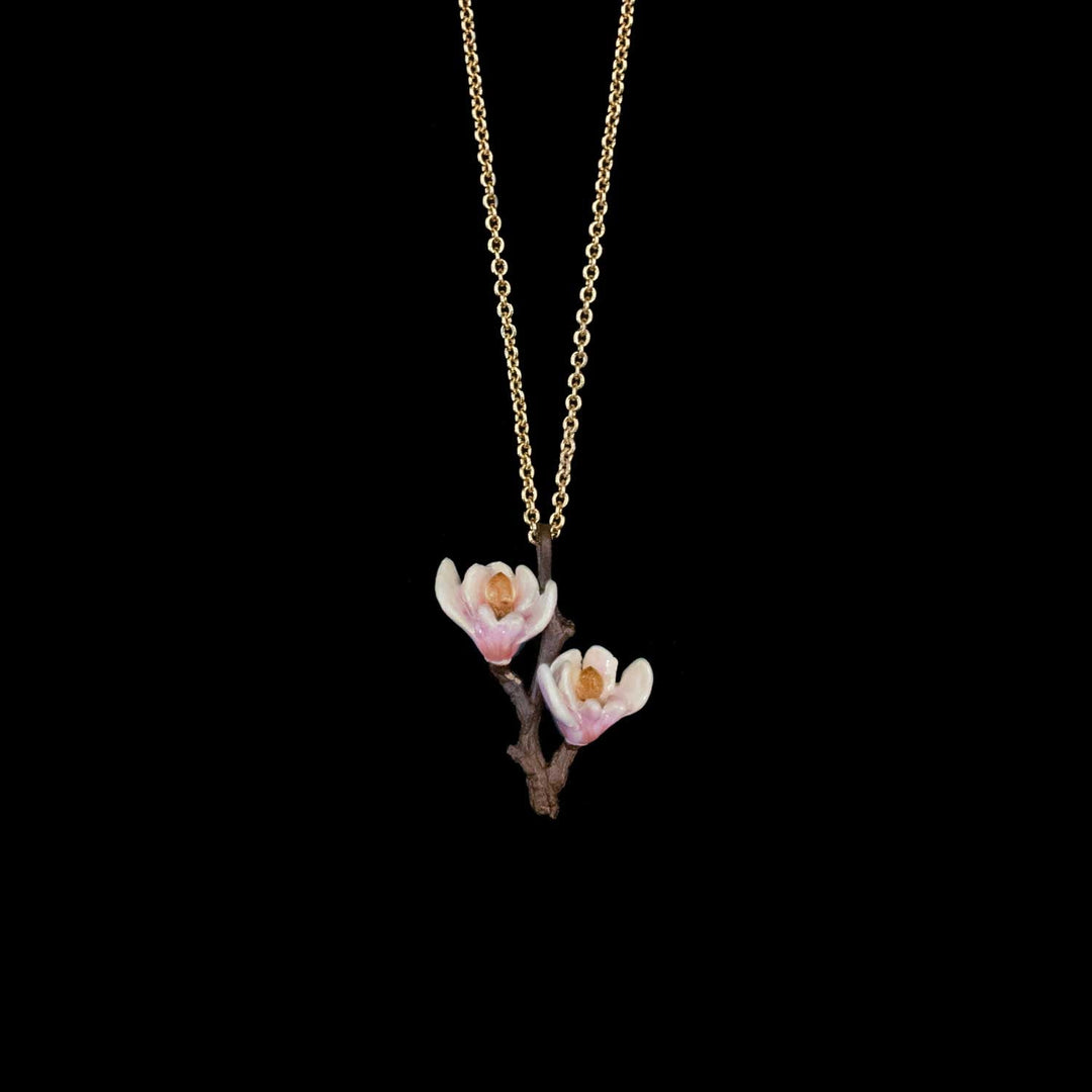 A delicate gold chain necklace featuring a pendant of two white magnolia blossoms with soft pink centers and a brown branch, set against a solid black background.