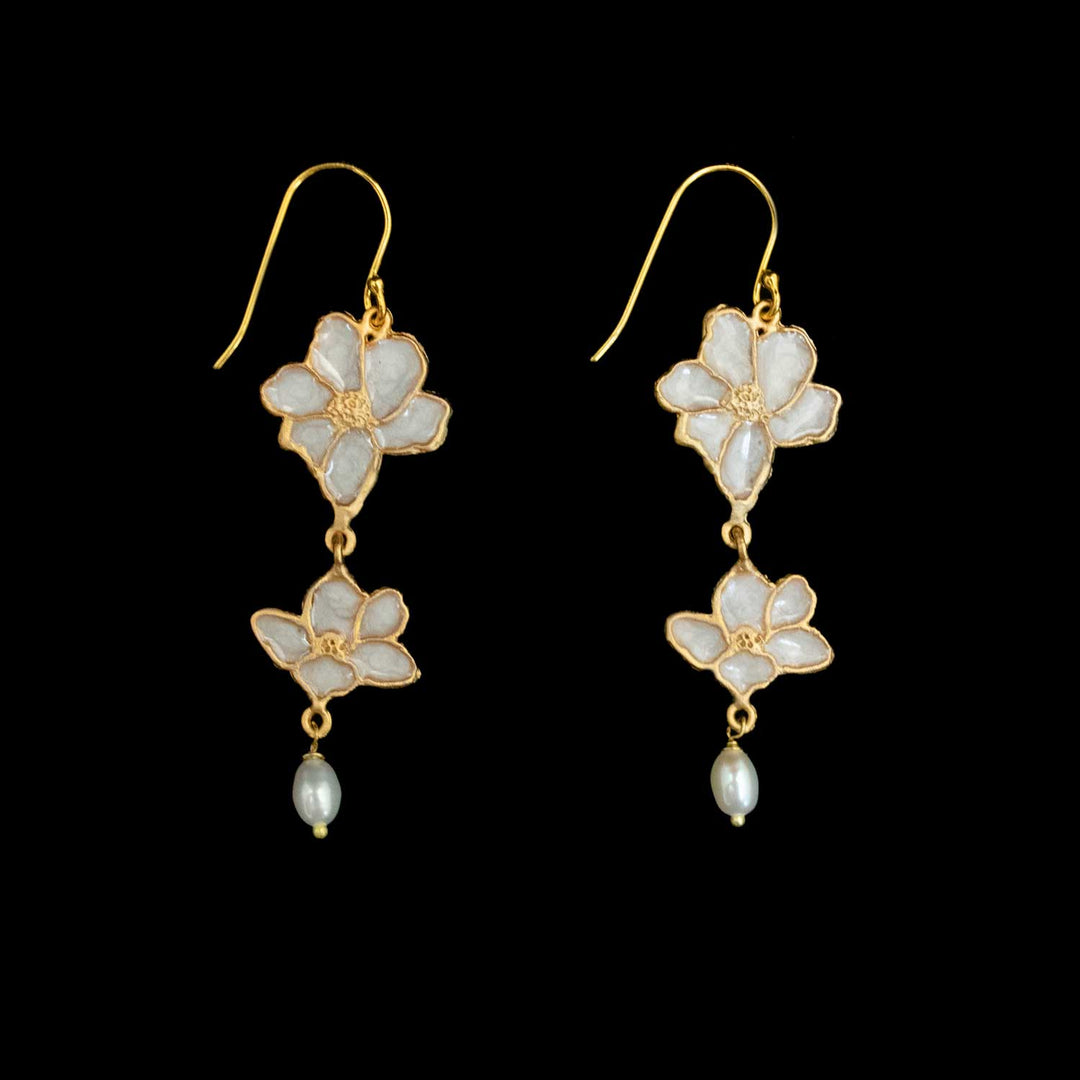 A pair of gold dangle earrings featuring white enamel flower charms and small pearl-like drops, set against a solid black background.