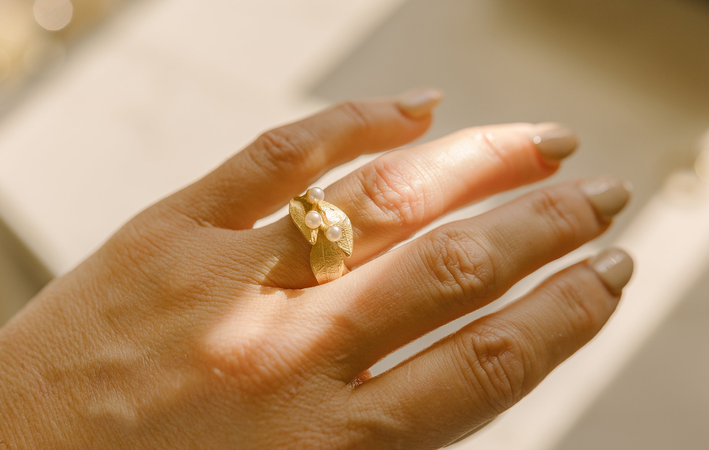A hand with neatly manicured nails wears a gold ring featuring small white pearls. The hand is lit by natural sunlight, casting soft shadows on the skin.