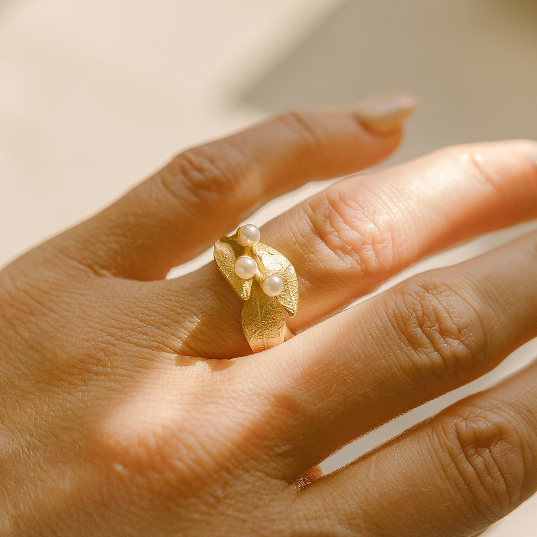 A hand with neatly manicured nails wearing a textured gold ring featuring three small pearl-like stones, shown in natural light.